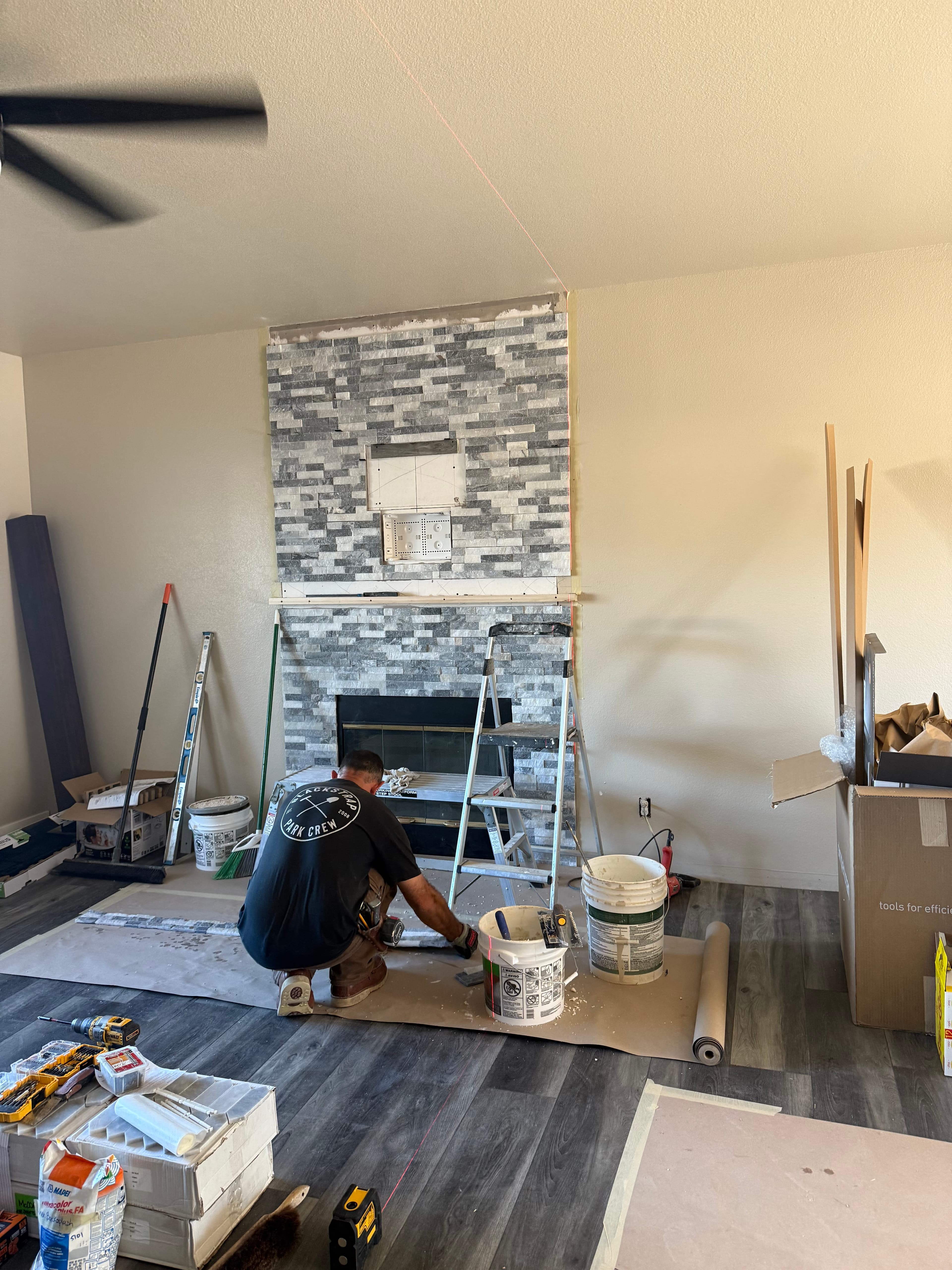 Man installing grey stone tiles on a fireplace wall with tools and buckets nearby.
